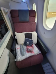 A first-class airplane seat with a maroon fabric upholstery is adjacent to two airplane windows. On the seat, a white pillow, a blanket in clear packaging, a pair of slippers, an amenity kit labeled with 'Japan Airlines', and a small red bag are neatly arranged.