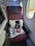 A first-class airplane seat with a maroon fabric upholstery is adjacent to two airplane windows. On the seat, a white pillow, a blanket in clear packaging, a pair of slippers, an amenity kit labeled with 'Japan Airlines', and a small red bag are neatly arranged.