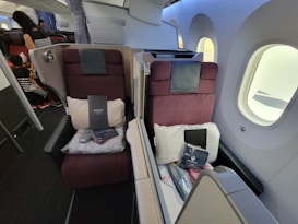 A luxurious airplane cabin featuring two burgundy seats with high backs, separated by a partition. Each seat is equipped with a pillow, a blanket, and a package labeled 'B.E.D.D.' The cabin has large windows, allowing natural light to brighten the space. In the aisle, a person and a child can be seen, indicating a comfortable and spacious environment.