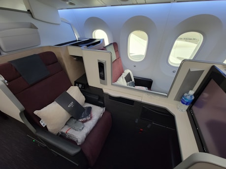 A luxurious airplane first-class cabin featuring a spacious seat that reclines into a bed. The seat is equipped with a pillow, blanket, and a provided amenity kit. Next to the seat, there is a water bottle and a screen for in-flight entertainment. The cabin is well-lit with natural light coming through the large windows. The interior design includes neutral and burgundy tones, creating an upscale atmosphere.