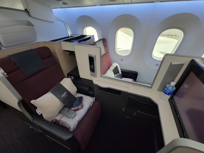 A luxurious airplane first-class cabin featuring a spacious seat that reclines into a bed. The seat is equipped with a pillow, blanket, and a provided amenity kit. Next to the seat, there is a water bottle and a screen for in-flight entertainment. The cabin is well-lit with natural light coming through the large windows. The interior design includes neutral and burgundy tones, creating an upscale atmosphere.