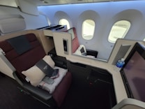 A luxurious airplane first-class cabin featuring a spacious seat that reclines into a bed. The seat is equipped with a pillow, blanket, and a provided amenity kit. Next to the seat, there is a water bottle and a screen for in-flight entertainment. The cabin is well-lit with natural light coming through the large windows. The interior design includes neutral and burgundy tones, creating an upscale atmosphere.