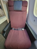A plush, reclining airplane seat upholstered in maroon fabric, featuring a seatbelt across it. The armrests are covered in dark leather or faux leather. A small part of the airplane cabin wall and a window are visible, enhancing the in-flight setting.