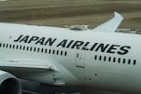 The image displays a large commercial aircraft on the tarmac, prominently featuring the Japan Airlines logo. The airplane is white with bold black lettering, and various ground service vehicles can be seen in the background.