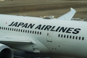 The image displays a large commercial aircraft on the tarmac, prominently featuring the Japan Airlines logo. The airplane is white with bold black lettering, and various ground service vehicles can be seen in the background.