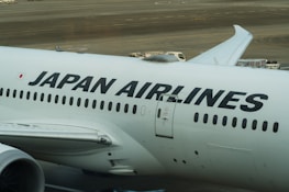 The image displays a large commercial aircraft on the tarmac, prominently featuring the Japan Airlines logo. The airplane is white with bold black lettering, and various ground service vehicles can be seen in the background.