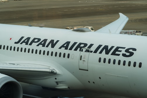 The image displays a large commercial aircraft on the tarmac, prominently featuring the Japan Airlines logo. The airplane is white with bold black lettering, and various ground service vehicles can be seen in the background.
