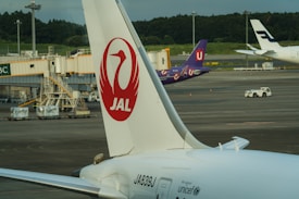 A close-up of a JAL airplane's tail with the company logo. In the background, there are airport facilities, including a boarding gate, and the tails of other airplanes. The scene is set on a tarmac with greenery in the distance and a clear sky.