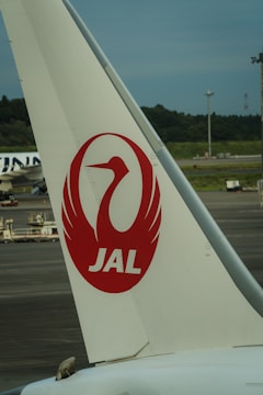 A close-up view of an airplane tail featuring the red logo of Japan Airlines, which includes a stylized crane. The background shows an airport runway with greenery and distant infrastructure.