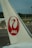 A close-up view of an airplane tail featuring the red logo of Japan Airlines, which includes a stylized crane. The background shows an airport runway with greenery and distant infrastructure.