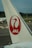 A close-up view of an airplane tail featuring a red logo of a stylized crane and the letters 'JAL' against a white background. The background includes airport tarmac, equipment, and distant trees under a cloudy sky.