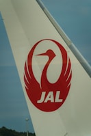 The image features the tail fin of an airplane with a white background. Prominently displayed on the tail is a red crane logo enclosed in a circle, along with the letters 'JAL' in bold red font below the logo. The sky in the background is a muted blue.