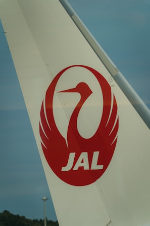 The image features the tail fin of an airplane with a white background. Prominently displayed on the tail is a red crane logo enclosed in a circle, along with the letters 'JAL' in bold red font below the logo. The sky in the background is a muted blue.