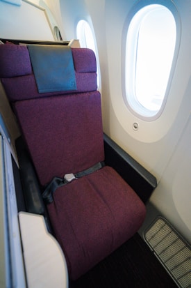 A comfortable-looking airplane seat is positioned next to two oval windows. The seat is upholstered in a dark maroon fabric and features a seatbelt across it. There is a leather headrest on the top of the seat, and the cabin appears modern and well-maintained.