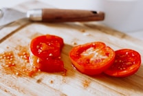 Slices of fresh red tomato are laid on a wooden cutting board. The board has a light, natural wood grain, and some tomato seeds and juice are visible around the slices. A metal kitchen utensil with a wooden handle is partially visible in the background.