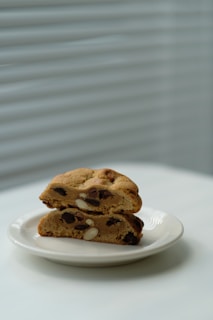 A pair of thick, freshly baked cookies with chocolate chips and nuts are stacked on a white plate. The background features soft, diffused light coming through white blinds, adding a serene and minimalistic feel.