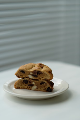 A pair of thick, freshly baked cookies with chocolate chips and nuts are stacked on a white plate. The background features soft, diffused light coming through white blinds, adding a serene and minimalistic feel.