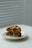A pair of thick, freshly baked cookies with chocolate chips and nuts are stacked on a white plate. The background features soft, diffused light coming through white blinds, adding a serene and minimalistic feel.