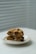 A pair of thick, freshly baked cookies with chocolate chips and nuts are stacked on a white plate. The background features soft, diffused light coming through white blinds, adding a serene and minimalistic feel.