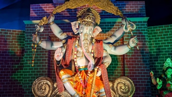 A detailed sculpture of a multi-armed deity with an elephant head, adorned in ornate jewelry and garments. The figure sits in a seated position against a backdrop of a brick pattern wall with vibrant colors. The deity holds various symbolic items and is decorated with a golden crown and intricate designs, exuding a sense of grandeur.
