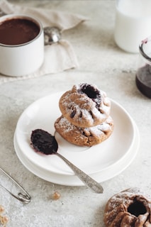 A neatly arranged dessert with two baked pastries topped with powdered sugar, placed on a white plate. Beside the pastries, a spoon holding a dark jam adds contrast. In the background, there is a jar of jam, a cup of dark beverage, possibly coffee or hot chocolate, and a white cloth napkin. The scene is set on a light, textured surface.