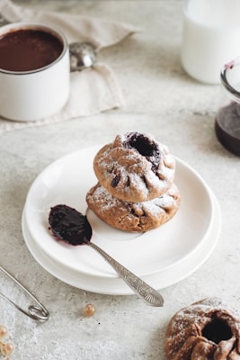 A neatly arranged dessert with two baked pastries topped with powdered sugar, placed on a white plate. Beside the pastries, a spoon holding a dark jam adds contrast. In the background, there is a jar of jam, a cup of dark beverage, possibly coffee or hot chocolate, and a white cloth napkin. The scene is set on a light, textured surface.