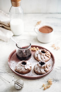 A light-colored ceramic tray holds three powdered sugar-dusted pastries surrounding a glass jar filled with dark liquid, which has a metal spoon. A white mug filled with a chocolate beverage, a bottle with a cork stopper containing milk, and a light-colored napkin are positioned in the background. Scattered around are some small branches of white currants and a tea infuser.