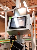 A manufacturing or industrial setting with machinery, including a control panel featuring a screen and keypad. The scene includes industrial lighting and parts of conveyor systems and structural pillars.