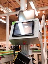 A manufacturing or industrial setting with machinery, including a control panel featuring a screen and keypad. The scene includes industrial lighting and parts of conveyor systems and structural pillars.