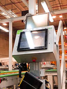 A manufacturing or industrial setting with machinery, including a control panel featuring a screen and keypad. The scene includes industrial lighting and parts of conveyor systems and structural pillars.