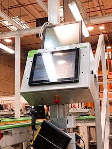 A manufacturing or industrial setting with machinery, including a control panel featuring a screen and keypad. The scene includes industrial lighting and parts of conveyor systems and structural pillars.