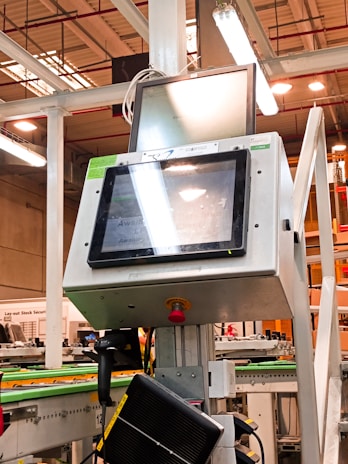 A manufacturing or industrial setting with machinery, including a control panel featuring a screen and keypad. The scene includes industrial lighting and parts of conveyor systems and structural pillars.
