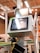 A manufacturing or industrial setting with machinery, including a control panel featuring a screen and keypad. The scene includes industrial lighting and parts of conveyor systems and structural pillars.