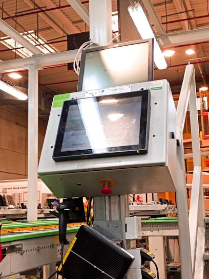 A manufacturing or industrial setting with machinery, including a control panel featuring a screen and keypad. The scene includes industrial lighting and parts of conveyor systems and structural pillars.