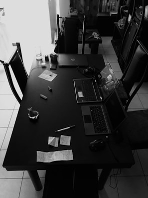 A black and white image displaying a table with two laptops, assorted stationery items such as a pen and sticky notes, along with a glass and some wrapped food items. The setting appears to be a casual workspace or dining area with chairs around the table.
