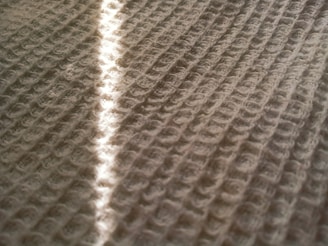 A close-up of a textured surface with a pattern of looped stitches, possibly a knitted or woven fabric. A streak of light runs diagonally across, highlighting the intricate design and adding depth to the composition.