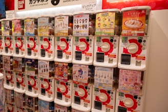 A row of gashapon capsule toy vending machines, each containing a different type of collectible toy. The machines are white with red accents, and there are various colorful posters and images on display on each machine advertising the toys inside.