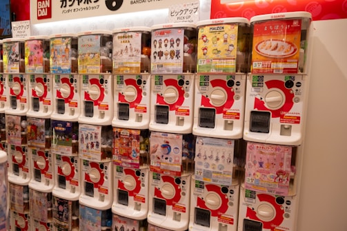 A row of gashapon capsule toy vending machines, each containing a different type of collectible toy. The machines are white with red accents, and there are various colorful posters and images on display on each machine advertising the toys inside.