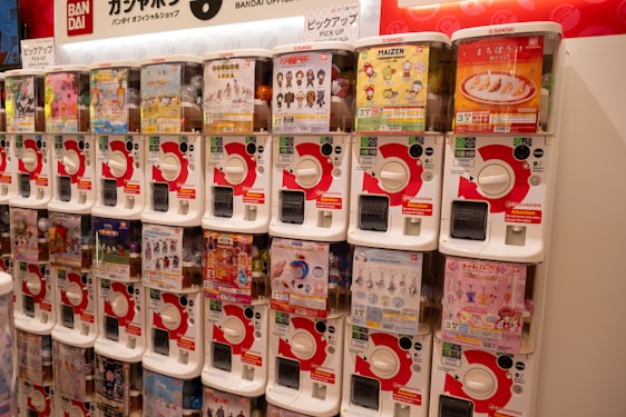 A row of gashapon capsule toy vending machines, each containing a different type of collectible toy. The machines are white with red accents, and there are various colorful posters and images on display on each machine advertising the toys inside.