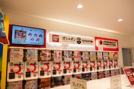 A row of gashapon capsule toy machines is lined up beneath a brightly lit interior with signs for Bandai and Japan Quality. There's a screen displaying promotional content, including toy images and text. Each vending machine has various toy options visible through transparent windows.