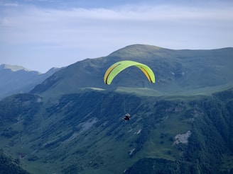 A paraglider with a bright yellow and black canopy soars over a vast, green mountainous landscape. The sky is clear with a soft blue hue, adding a serene backdrop to the scene. The rugged terrain of the mountains below features patches of forest and rocky outcrops.