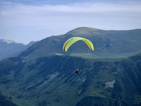 A paraglider with a bright yellow and black canopy soars over a vast, green mountainous landscape. The sky is clear with a soft blue hue, adding a serene backdrop to the scene. The rugged terrain of the mountains below features patches of forest and rocky outcrops.