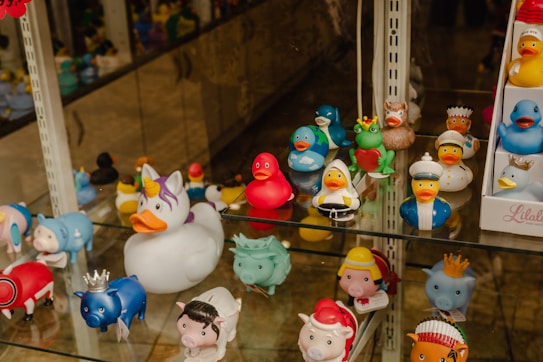 A collection of rubber duck toys displayed on shelves, each designed with unique themes ranging from a unicorn to a sailor and various animal costumes. The ducks are brightly colored and arranged in a whimsical fashion.