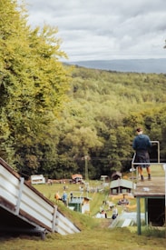 A person stands on a platform overlooking a grassy slope leading to a small rural area. The scene is surrounded by dense green trees under a cloudy sky. In the distance, several small buildings and scattered people are visible, giving the impression of a community gathering or event.