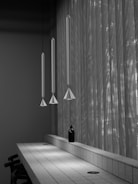 A minimalist interior features three elongated pendant lamps hanging above a sleek, tiled counter. A small black bottle is placed prominently on the counter, while sunlight filters through sheer, textured curtains, casting subtle shadows.
