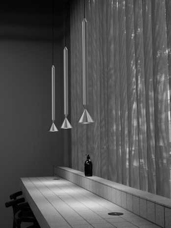 A minimalist interior features three elongated pendant lamps hanging above a sleek, tiled counter. A small black bottle is placed prominently on the counter, while sunlight filters through sheer, textured curtains, casting subtle shadows.