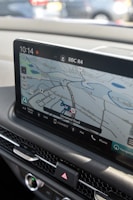 A car's dashboard displaying a digital navigation system. The screen shows a map with various road names and a highlighted route. The time, radio station (BBC R4), and various control icons are visible. Below the screen are car control buttons, including the hazard light switch.