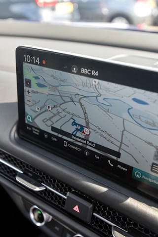 A car's dashboard displaying a digital navigation system. The screen shows a map with various road names and a highlighted route. The time, radio station (BBC R4), and various control icons are visible. Below the screen are car control buttons, including the hazard light switch.