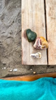 Several uniquely shaped stones rest on a wooden surface, with a sandy texture visible in the background. A vibrant turquoise fabric is seen at the lower edge, adding contrast to the natural colors of the stones and wood.