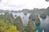 Lush, green cone-shaped islands rise from tropical turquoise waters, surrounded by dense vegetation. Bright skies with scattered clouds form the backdrop. The landscape suggests a serene, remote paradise, with calm waters and a natural, untouched environment.